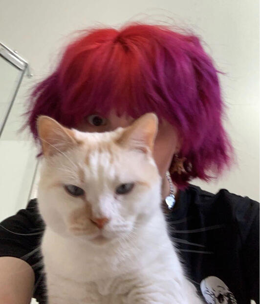 A selfie of a young person with short wavy red and purple hair. Their face is covered by a mildly disgruntled but curious white cat with light orange stripes.